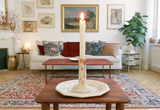 A lit white taper candle in a vintage candle holder sitting on a wooden table in a Scandinavian living room