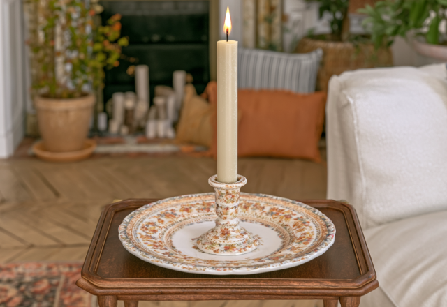 A vintage candle holder with a lit taper candle resting on a wooden table in a living room.