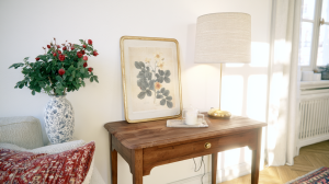Vintage console table styling with botanical art, a brass lamp, and a vase of red roses in a bright living room.