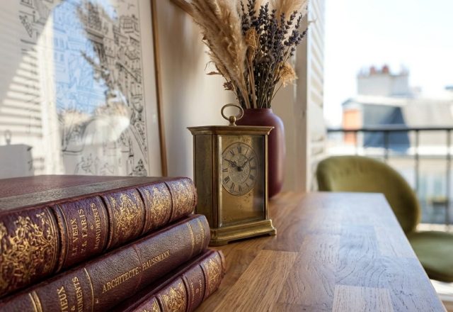 Vintage desk decor featuring leather bound books, a brass clock, and framed art on a wooden surface.