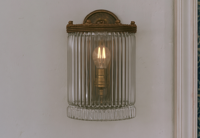 A vintage glass wall sconce with a ribbed shade and warm filament bulb mounted on a textured white wall.