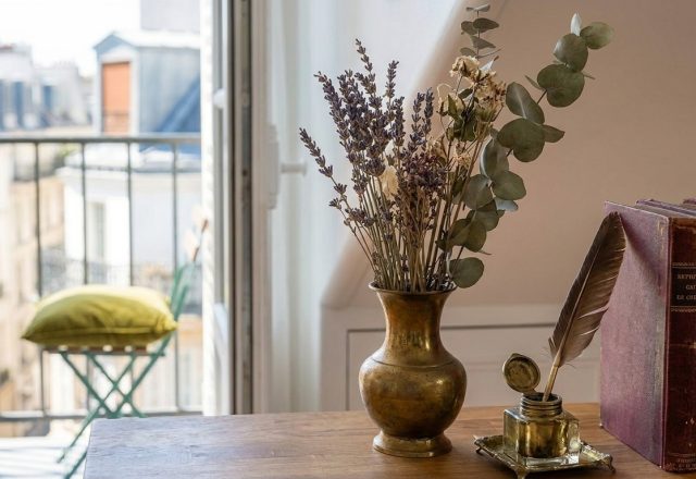 Vintage home office styling featuring brass vase with dried lavender, inkwell with quill, and antique books next to a balcony window.