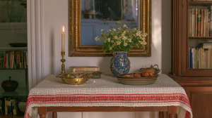 Vintage tabletop decor featuring an antique mirror, brass candle holder, and blue ceramic vase with daisies on a red-striped runner.