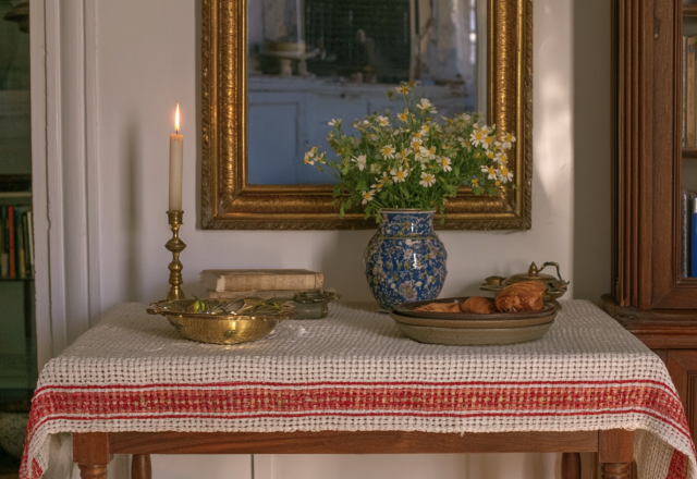 Vintage tabletop decor featuring an antique mirror, brass candle holder, and blue ceramic vase with daisies on a red-striped runner.