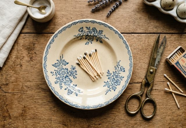 Vintage tabletop organization featuring a blue and white patterned plate, matches, and antique scissors on a rustic wooden table.