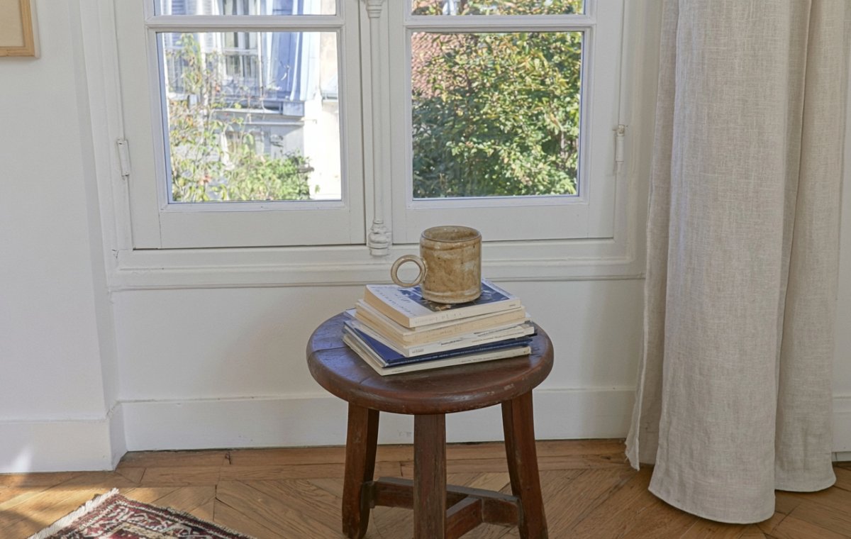 A vintage wooden stool holding stacked books and a ceramic mug near a bright window.