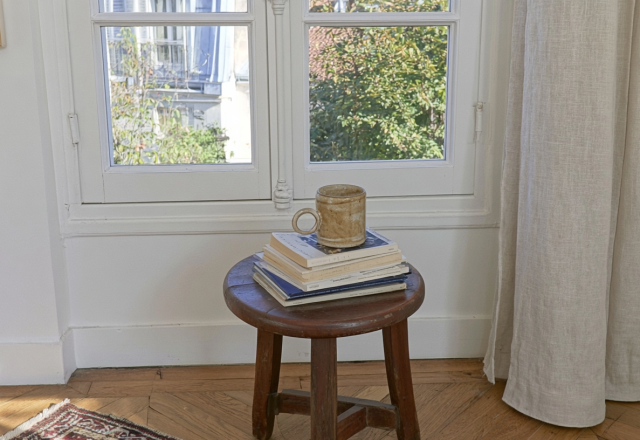 A vintage wooden stool holding stacked books and a ceramic mug near a bright window.