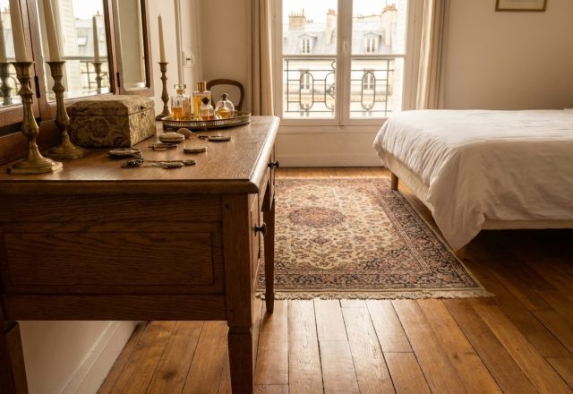 Vintage wooden vanity with brass candlesticks and perfume bottles beside a classic white bed in a bright bedroom