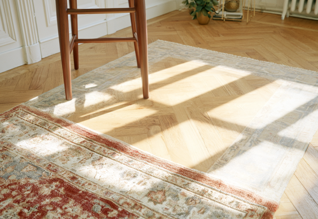 Vintage wool rug layering on herringbone wood floors in a sunlit living room with a mid-century stool.