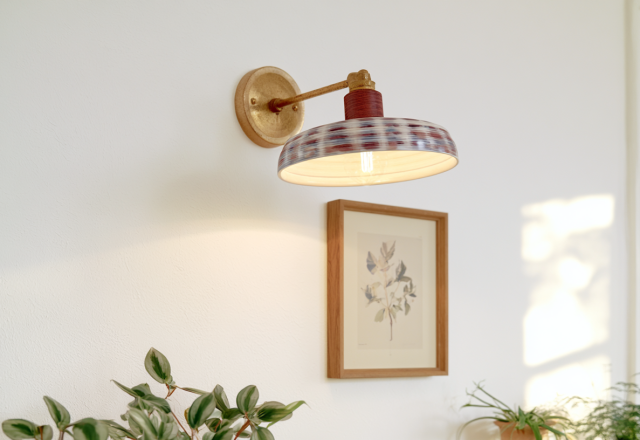 A decorative patterned wall sconce lighting fixture mounted above framed botanical art and green plants.