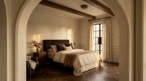 A cozy warm minimalist bedroom featuring earthy tones, rustic beams, and herringbone flooring viewed through an arched doorway.