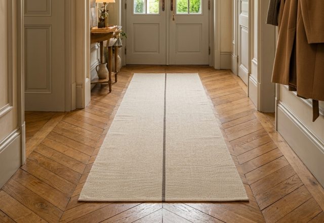 A cream hallway runner rug with a dark stripe on chevron wood flooring in a traditional home.