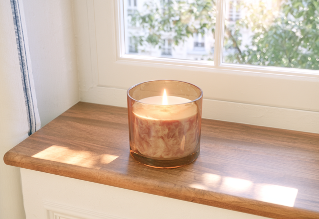 An amber glass candle burning on a wooden window sill with daylight streaming through the panes.