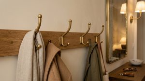 A minimalist wooden coat rack with brass hooks mounted on a beige wall holding winter coats and scarves.