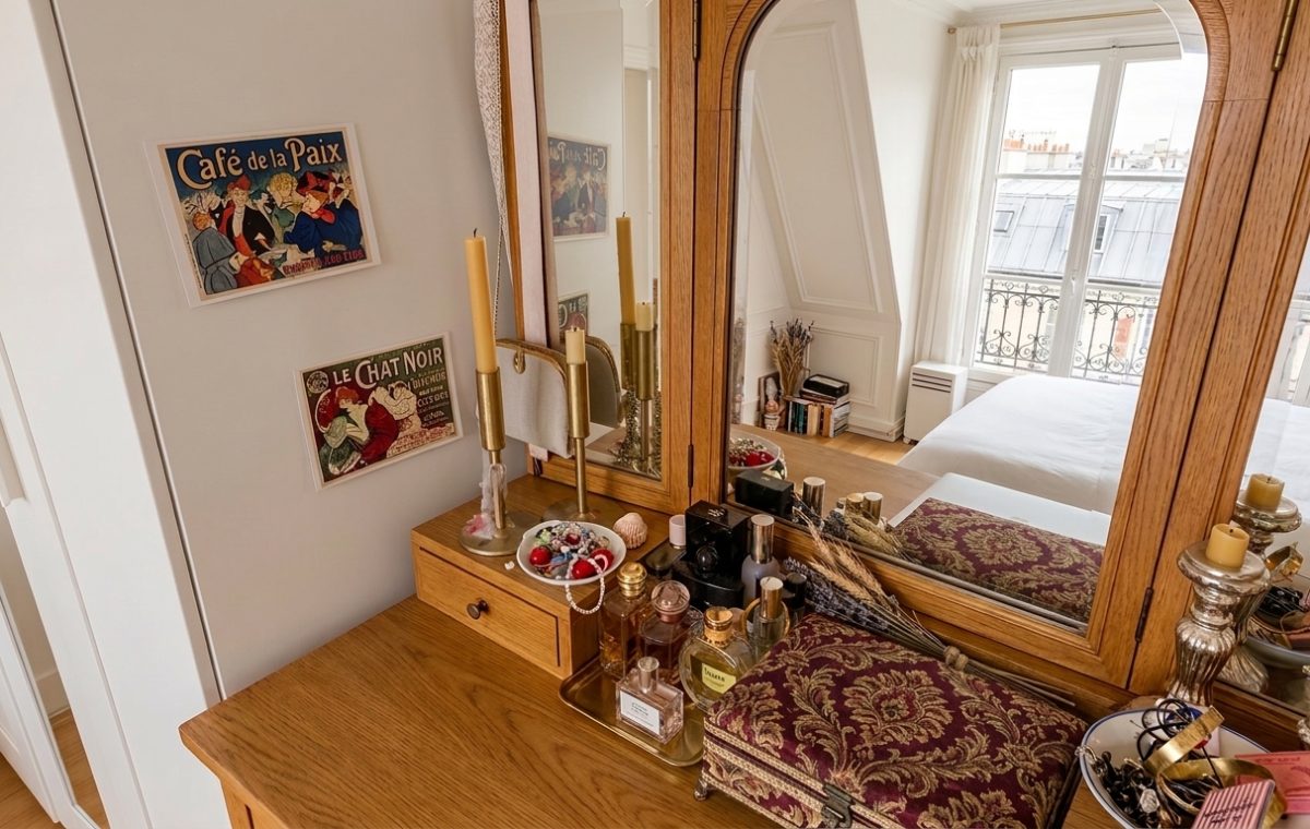 A wooden vanity mirror styled with vintage perfume bottles, brass candlesticks, and jewelry on a dressing table in a bright bedroom.