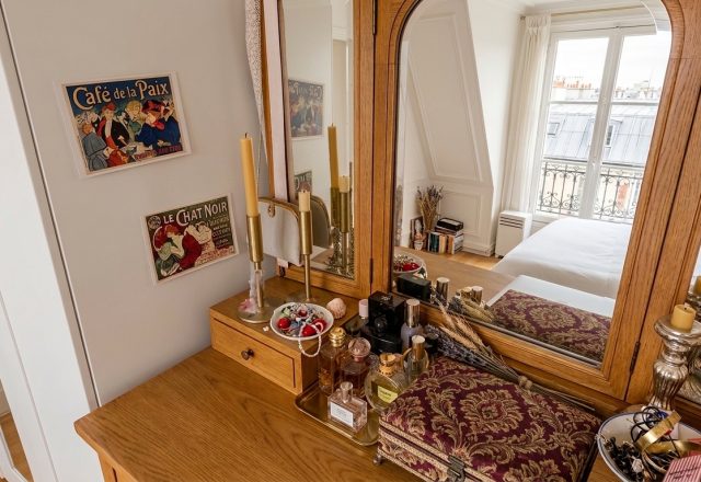 A wooden vanity mirror styled with vintage perfume bottles, brass candlesticks, and jewelry on a dressing table in a bright bedroom.