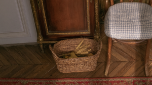 A rustic woven storage basket filled with dried wheat sits on hardwood parquet flooring next to an antique cabinet and upholstered chair.